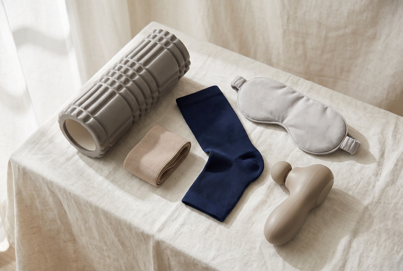 Everyday wellness essentials laid out together: a foam roller, a resistance band, a compression sleeve, a sleep mask and a handheld massager